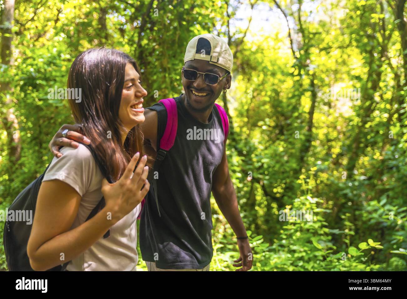Coppia multi etnica felice che si diverte a fare trekking in una foresta verde, indossando zaini e abiti casual Foto Stock