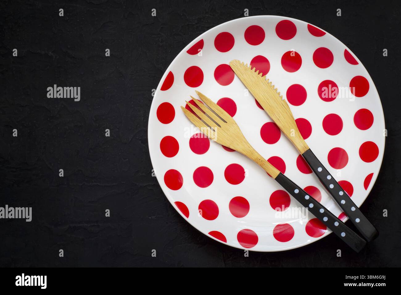 Tavolo con coltello in bambù, forchetta e piatto con puntini di polka su sfondo scuro Foto Stock