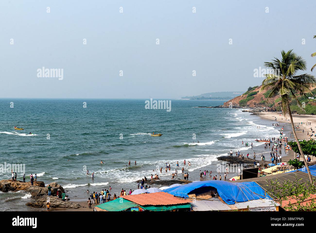 A Anjuna Beach a Goa, India, le onde dolci si infrangono su aspre formazioni rocciose lungo la costa, mentre numerosi visitatori si impegnano in attività sulla spiaggia Foto Stock