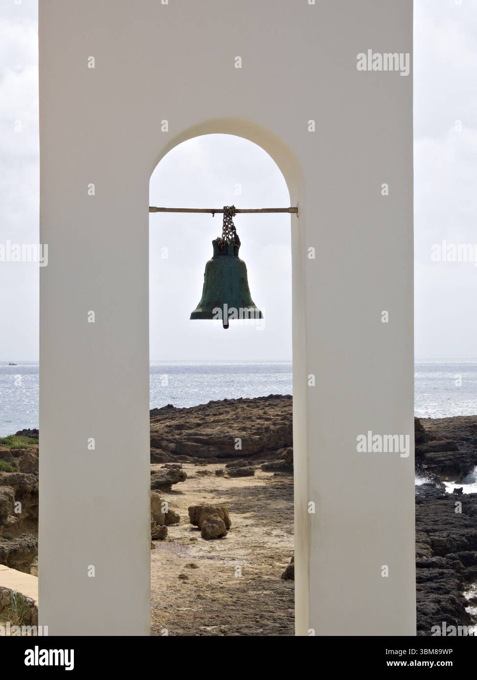 Piccola parete bianca con campana singola, incorniciata da costa frastagliata e cielo nuvoloso, Grecia. Foto Stock