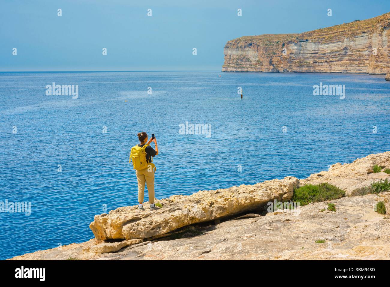 Donna viaggiatrice, vista posteriore di una giovane donna che indossa uno zaino giallo che fotografa un tratto della costa mediterranea di Malta Foto Stock