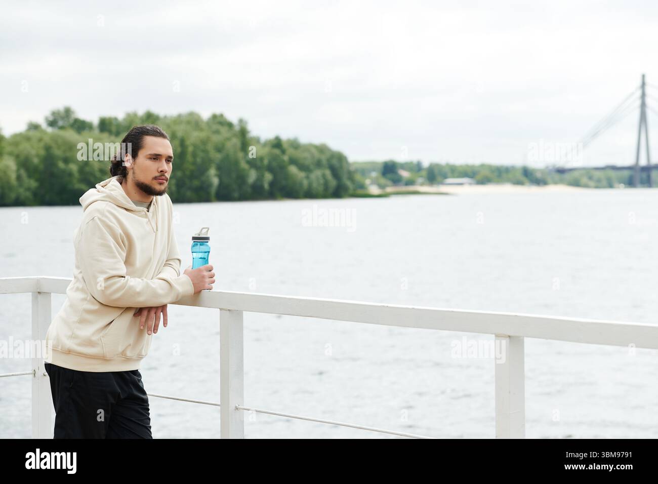 Impegnato in attività all'aperto, un uomo in forma riposa vicino all'acqua con una bevanda in mano. Foto Stock
