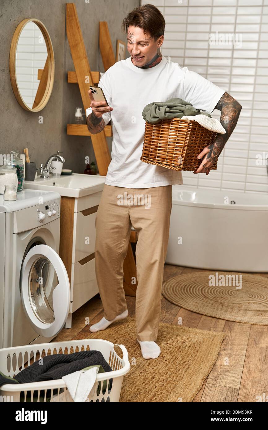 Il bel tatuato bilancia i compiti di lavanderia mentre si interagisce con il telefono a casa. Foto Stock