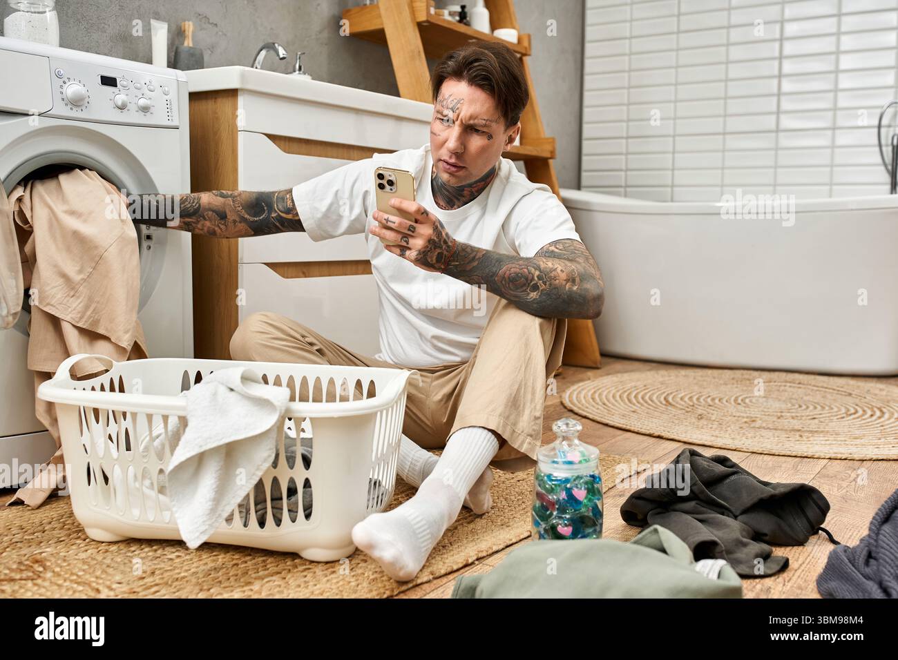 Un giovane tatuato si siede comodamente a casa, controllando il telefono mentre si fa il bucato. Foto Stock