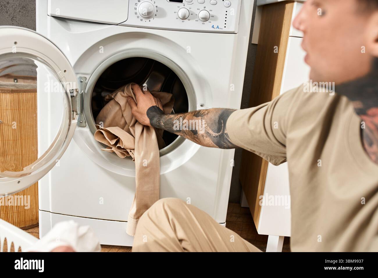 Un giovane con i tatuaggi mette i vestiti in lavatrice mentre si rilassa a casa. Foto Stock
