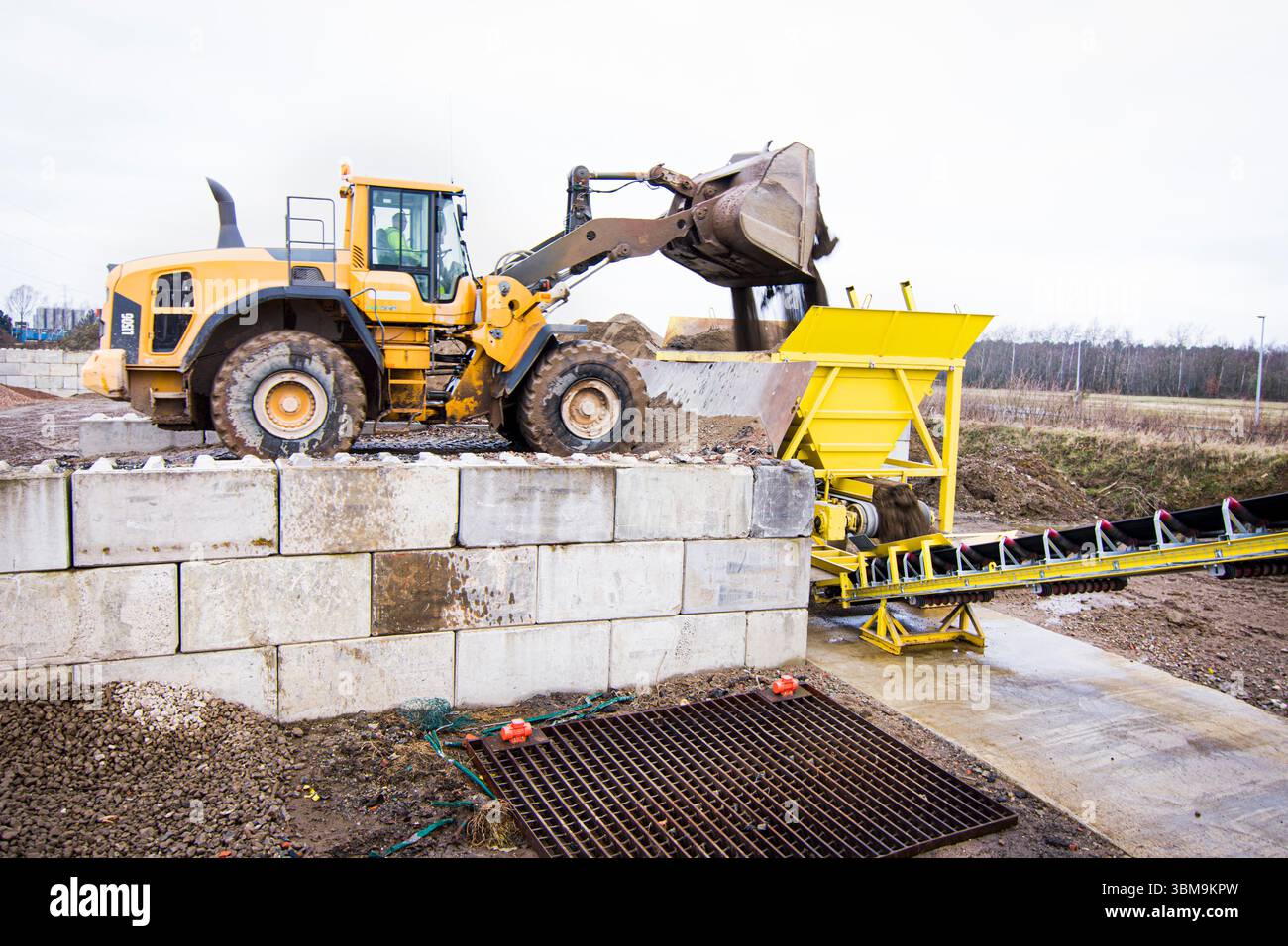 Caricatore frontale per impieghi gravosi che scarica la ghiaia in una tramoggia del convogliatore gialla in un cantiere industriale. Macchine movimento terra in azione su un impianto Foto Stock