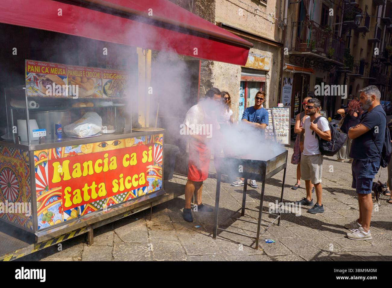 Questa immagine cattura la vibrante energia del mercato di Ballarò a Palermo, in Sicilia. Un chiosco di cibo con una tenda rossa serve come punto focale, dove un uomo gri Foto Stock