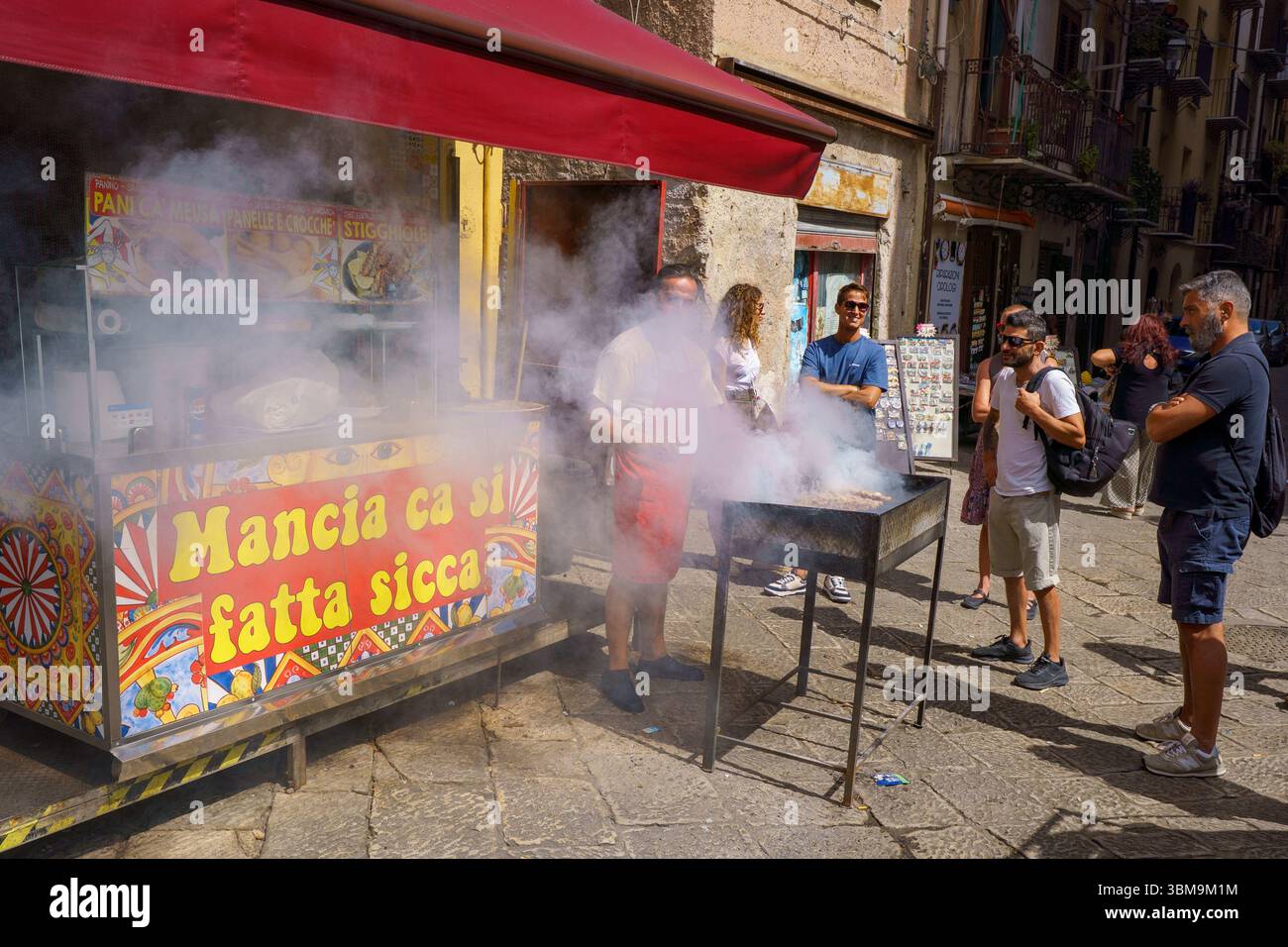 Questa immagine cattura la vibrante energia del mercato di Ballarò a Palermo, in Sicilia. Un chiosco di cibo con una tenda rossa serve come punto focale, dove un uomo gri Foto Stock