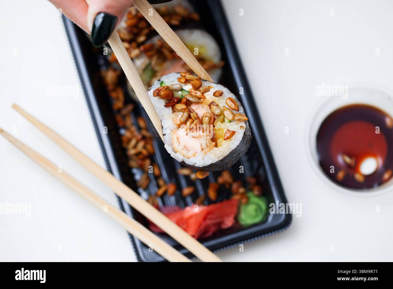 Sushi roll tenuto da bacchette. Cibo asiatico con riso e verdure. Salsa di soia e tavolo in legno sullo sfondo. Vassoio da asporto nero di sushi. Foto Stock