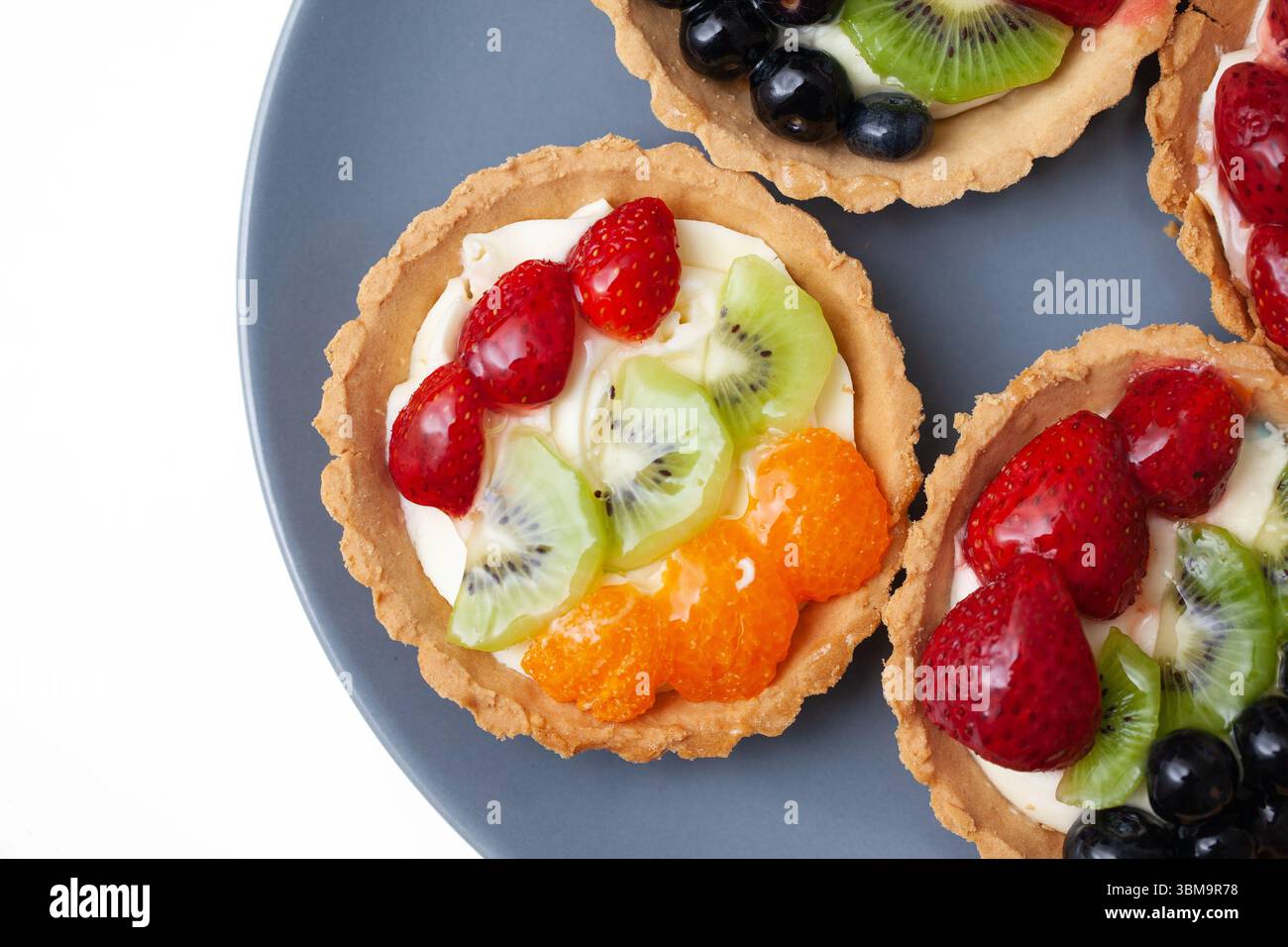 Crostate di pasticceria con frutta fresca. Mini torte colorate con kiwi fragola arancia e uva. Dessert dolce su piatto blu. Ingredienti naturali. Foto Stock