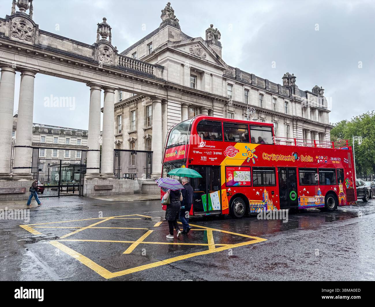 Autobus a due piani City Sightseeing a Dublino durante una giornata di pioggia. I pedoni con ombrelloni camminano sulla strada bagnata vicino a un grande edificio storico. Foto Stock