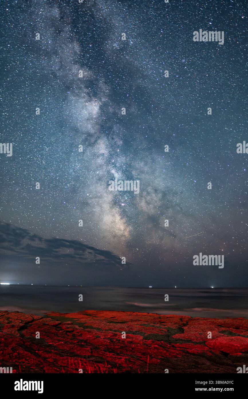 La via Lattea si innalza sopra un mare calmo e rocce costiere illuminate di rosso, mescolando meraviglia cosmica con le texture elementari della Terra in un tranquillo paesaggio notturno. Foto Stock