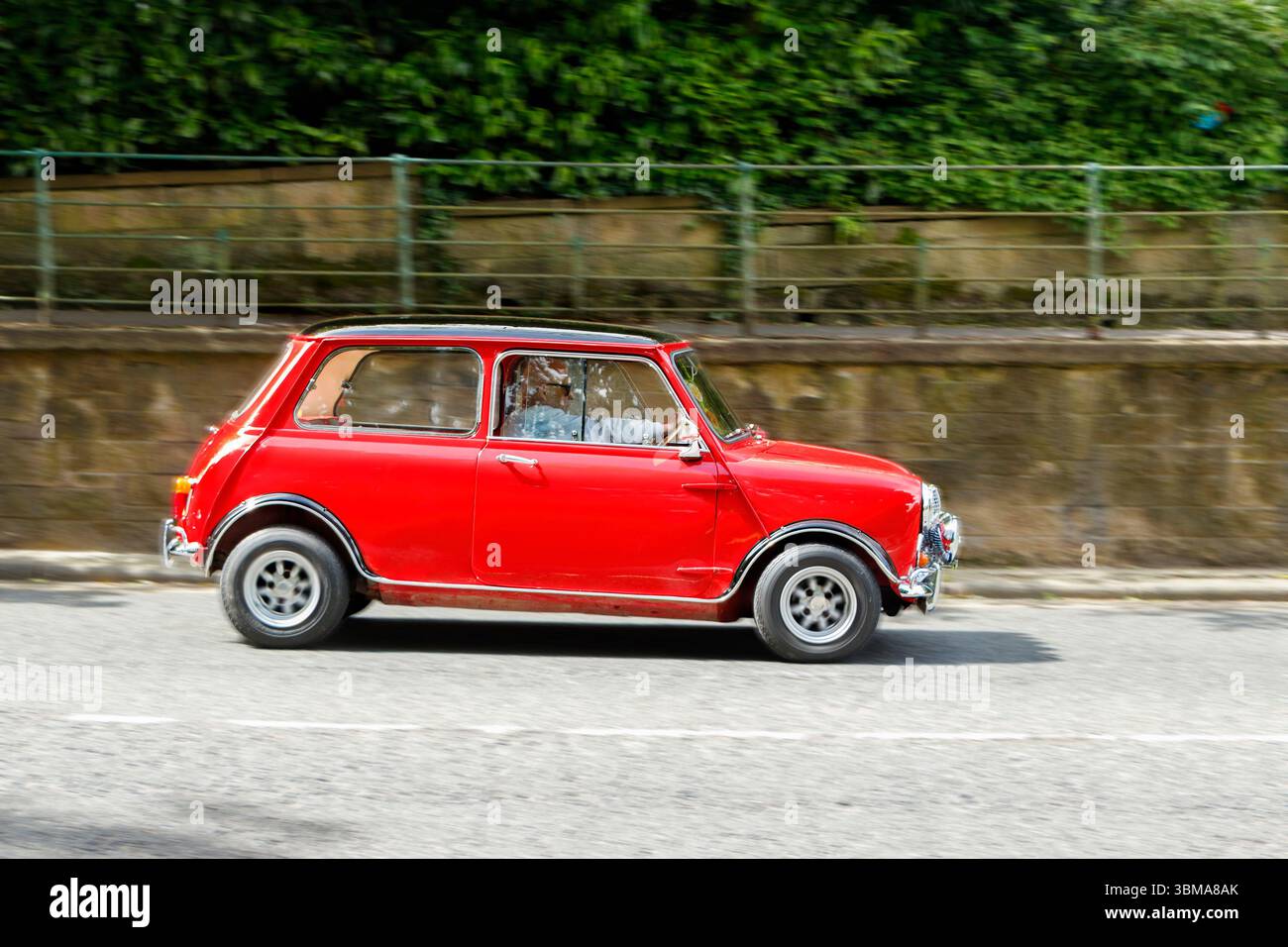 Mini Cooper rossa classica che guida lungo una strada con un muro di pietra e vegetazione sullo sfondo Foto Stock
