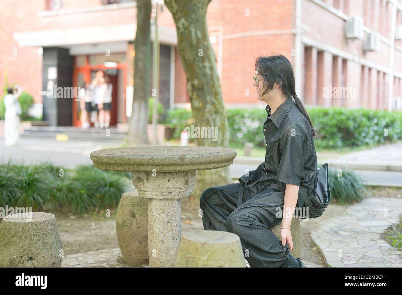 Un giovane cinese di vent'anni, con gli occhiali, siede tranquillamente su una panchina di legno nel campus, godendosi la brezza, circondato da alberi in fiore e m Foto Stock Un giovane cinese di vent'anni, con gli occhiali, siede tranquillamente su una panchina di legno nel campus, godendosi la brezza, circondato da alberi in fiore e m Foto Stock