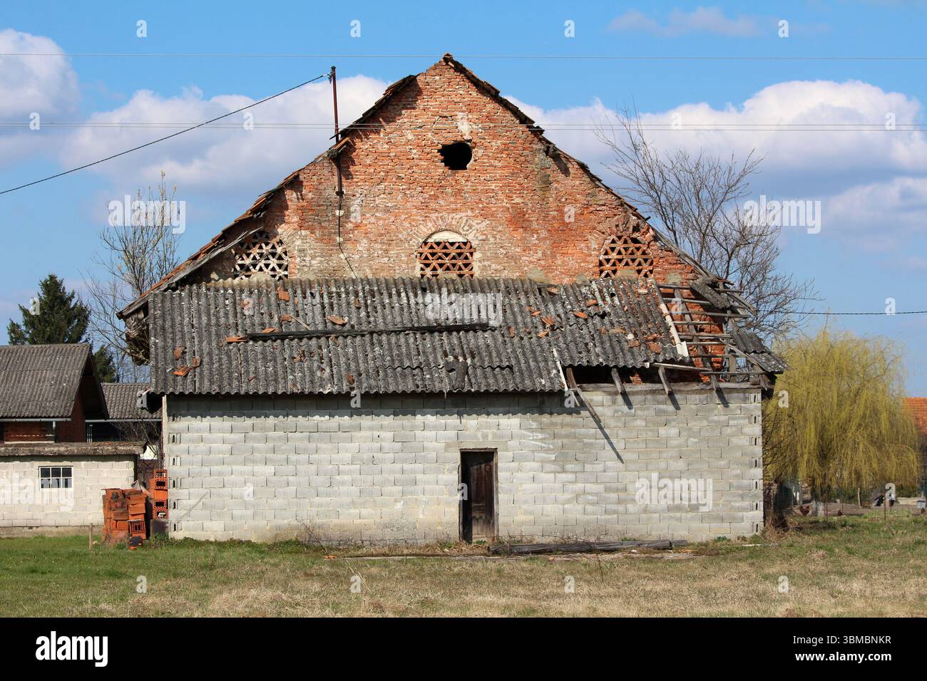 Vecchio fienile costruito in mattoni rossi e blocchi di cemento grigio, caratterizzato da un tetto in piastrelle parzialmente crollato e sezioni mancanti, sorge in una zona rurale erbosa Foto Stock