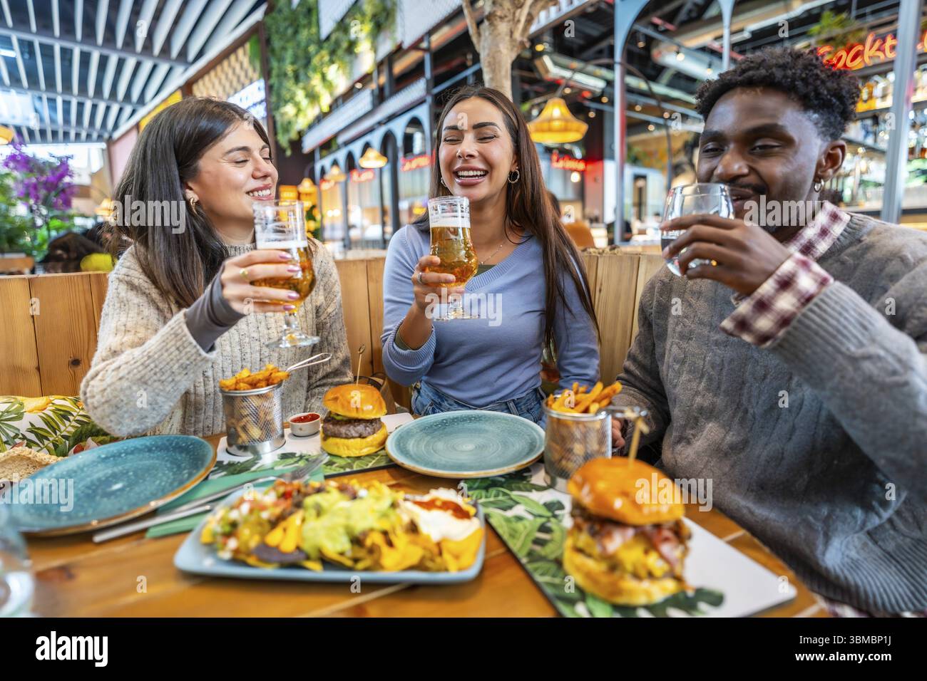 Amici multietnici felici si divertono insieme, mangiando hamburger, nachos e patatine fritte e bevendo birra al ristorante Foto Stock