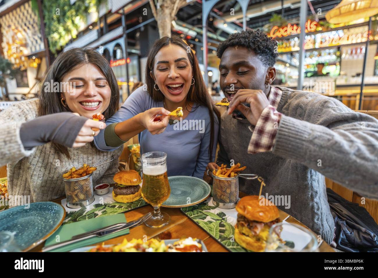 Tre allegri amici multietnici che condividono nachos e hamburger, gustando un pasto insieme in un ristorante Foto Stock