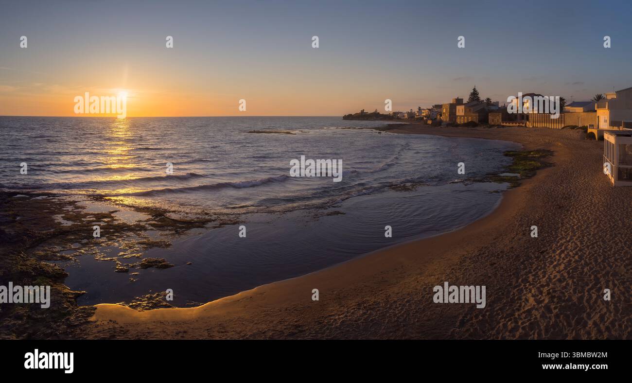 Il sole dorato si tuffa all'orizzonte a Punta secca, riflettendo sulle acque tranquille e proiettando un caldo bagliore sulla costa, in Sicilia, Italia Foto Stock