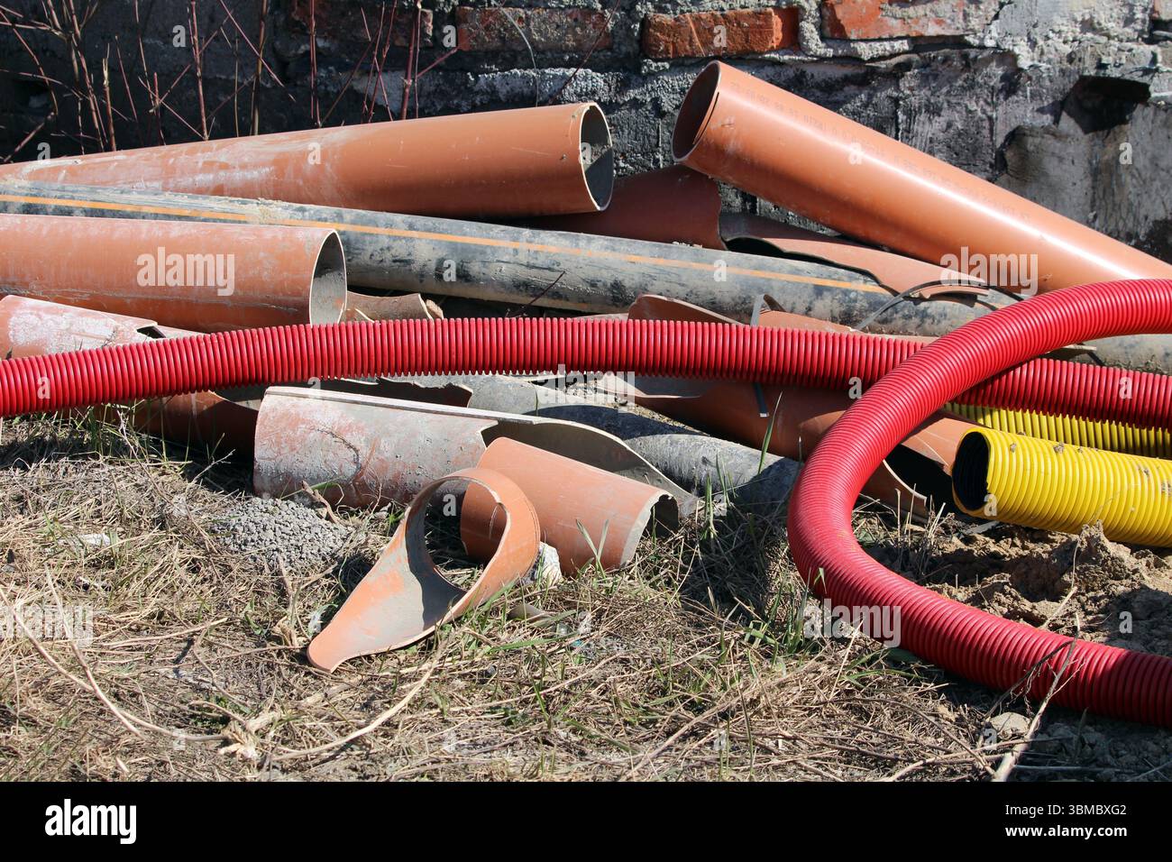 Pile miste di tubi di plastica e metallo arancioni, neri, rossi e gialli, alcune ondulate, giacciono su erba secca accanto a una parete di mattoni, indicando la costruzione Foto Stock
