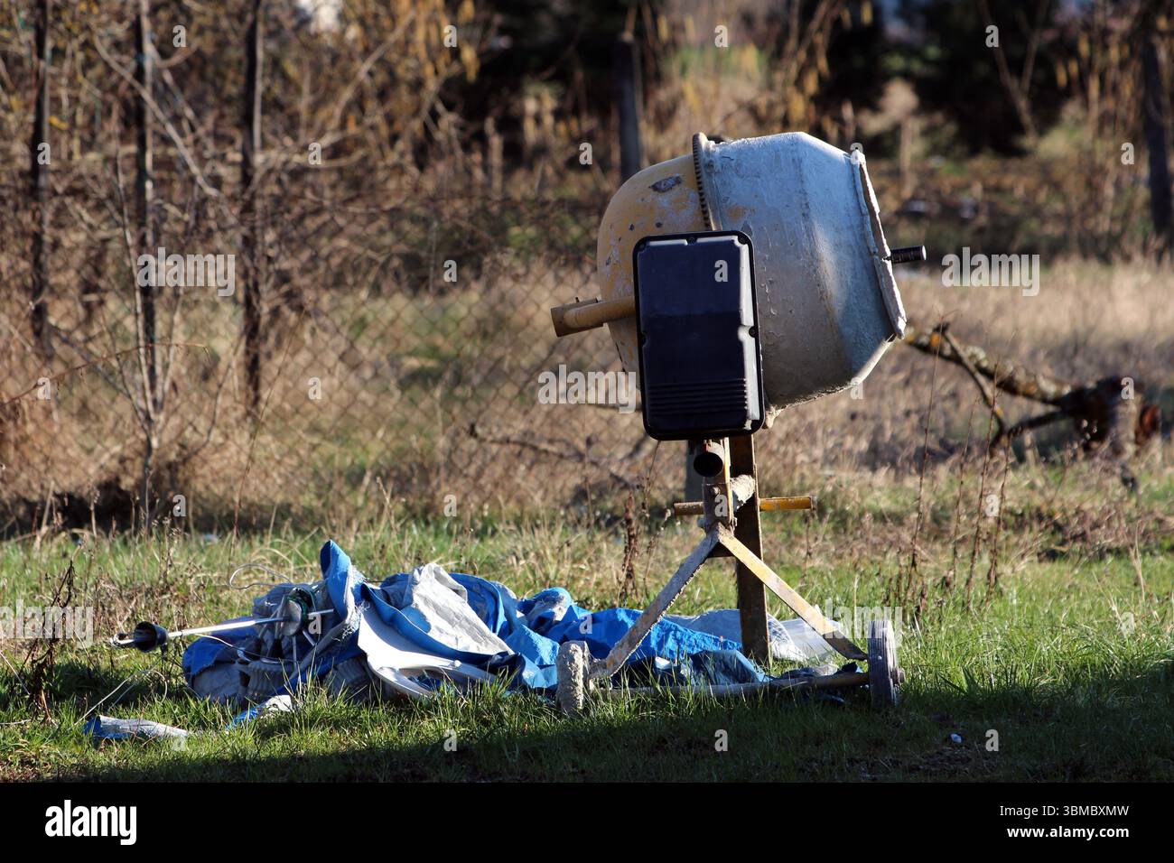 La betoniera portatile si trova sulle gambe di un treppiede accanto a un telo blu e bianco deformato, su erba verde con una recinzione metallica e vegetazione secca Foto Stock