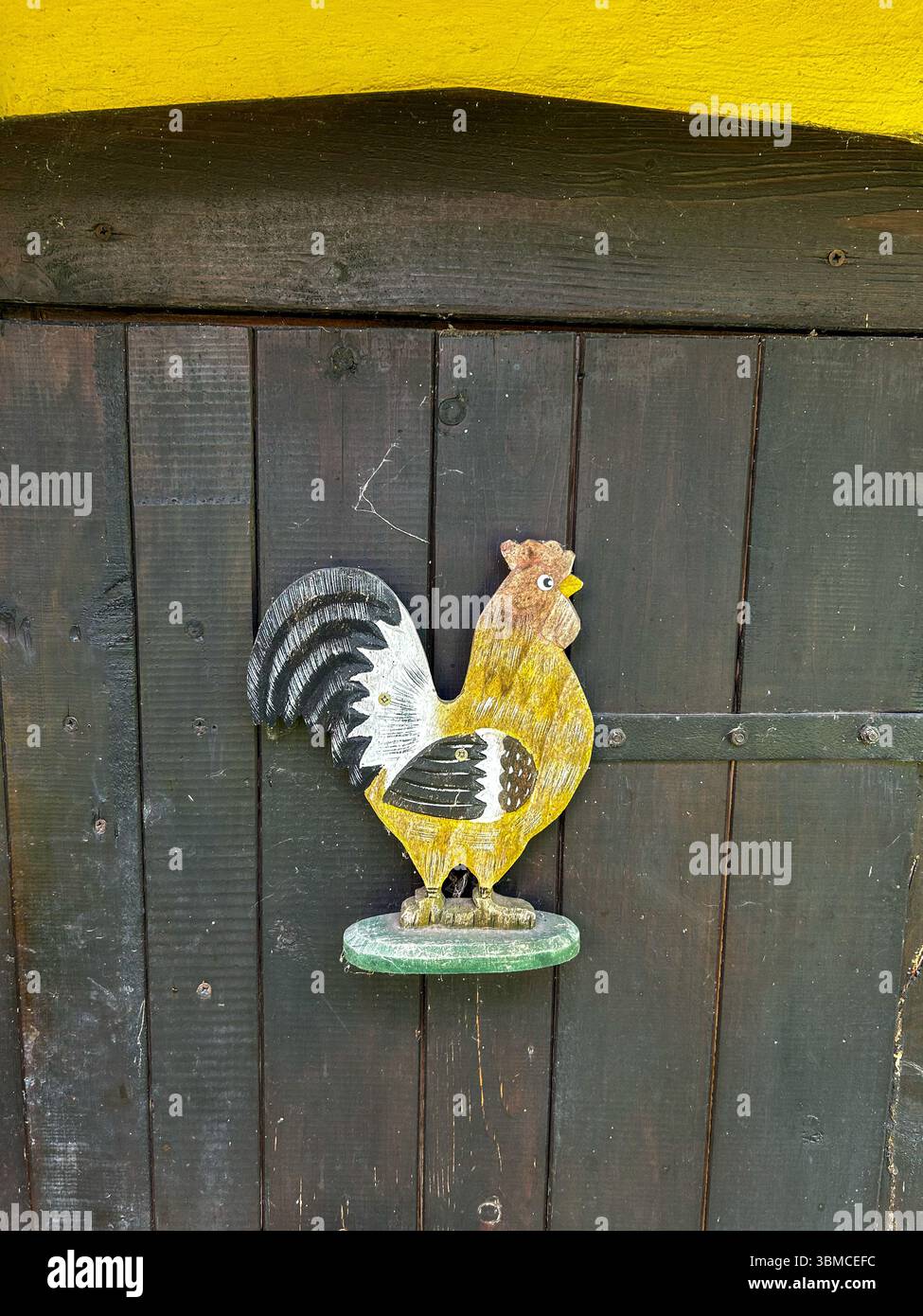 Decorazioni in legno di gallo dipinte a mano montate su una porta di fienile di legno scuro, che aggiungono un tocco di fascino rurale e carattere vintage al set di campagna Foto Stock