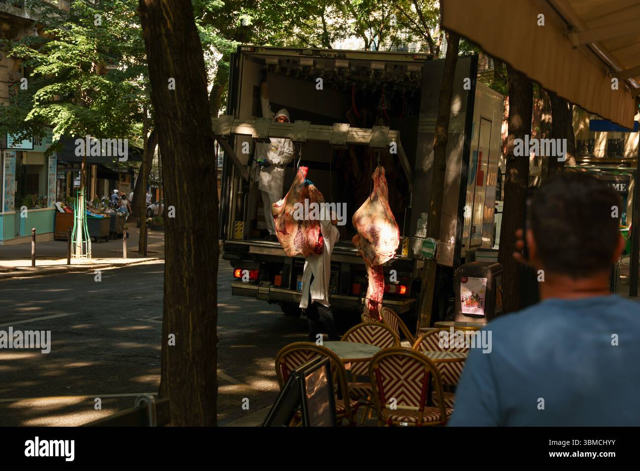 Due macellai in cappotti bianchi scaricano con cura grandi lati di manzo da un camion refrigerato parcheggiato in una soleggiata strada parigina. Foto Stock