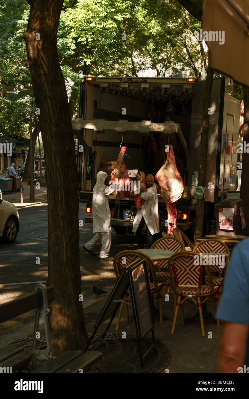 Due macellai con indumenti protettivi bianchi scaricano con cura grandi lati di manzo da un camion refrigerato parcheggiato in una strada parigina. Foto Stock