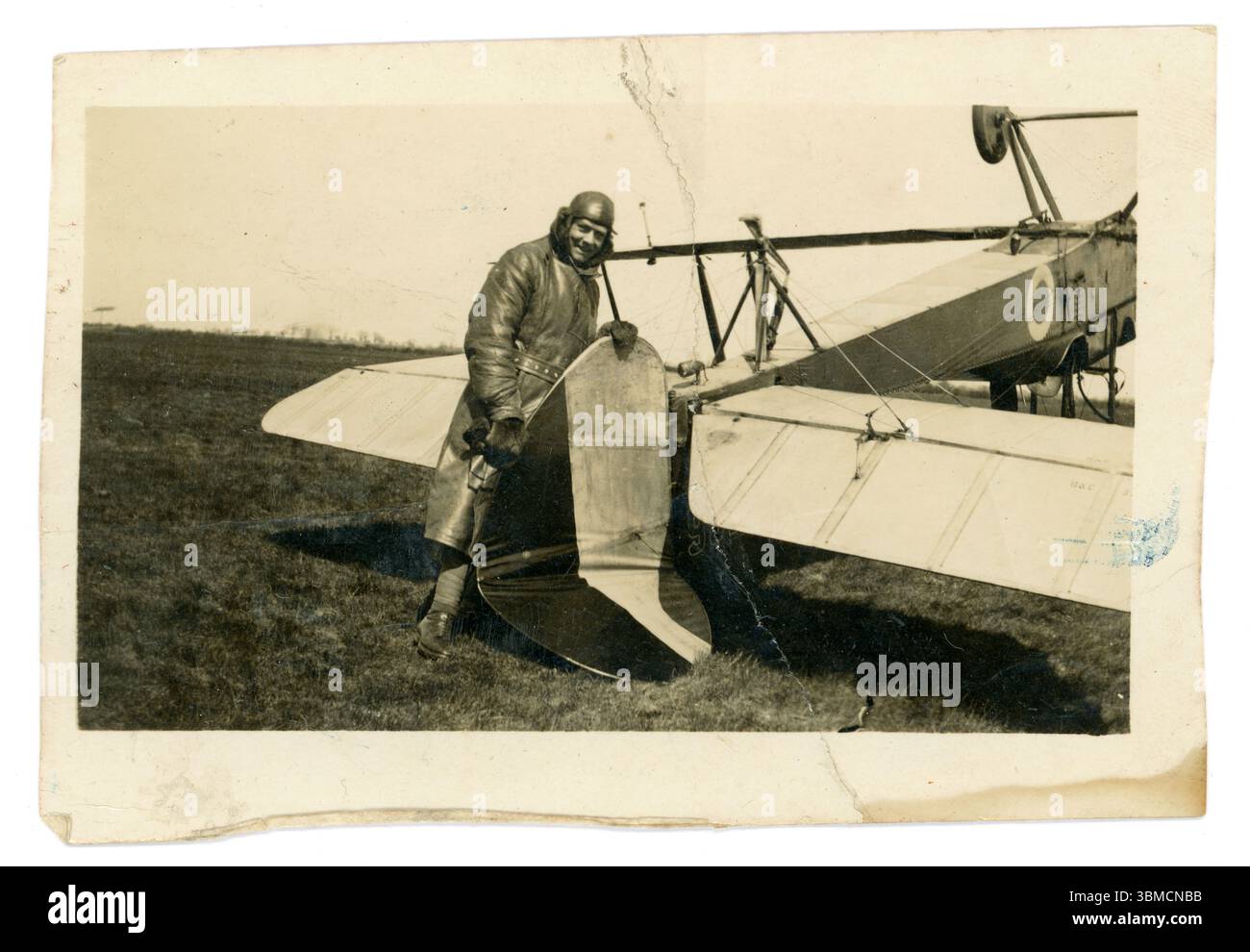Aereo originale dell'era WW1 con un pilota dall'aspetto allegro, possibilmente del Royal Flying Corps (RFC) in equipaggiamento volante, tra cui un lungo trench in pelle, un berretto e guanti in pelle, in piedi accanto al suo aereo capovolto, che si è ribaltato e si è schiantato: Tiene in mano la coda rotta e grattugia. 1914-1918 Foto Stock