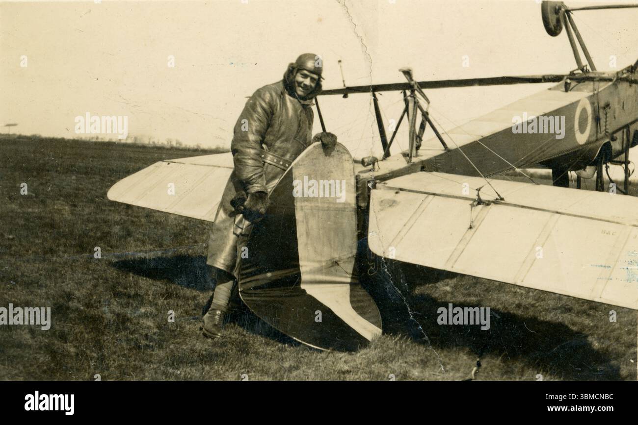 Aereo originale dell'era WW1 con un pilota dall'aspetto allegro, possibilmente del Royal Flying Corps (RFC) in equipaggiamento volante, tra cui un lungo trench in pelle, un berretto e guanti in pelle, in piedi accanto al suo aereo capovolto, che si è ribaltato e si è schiantato: Tiene in mano la coda rotta e grattugia. 1914-1918 Foto Stock