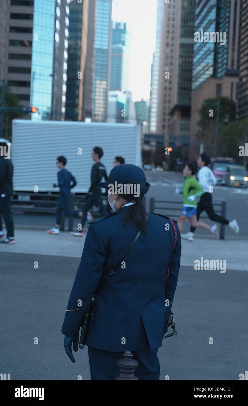 Ufficiale di sicurezza in uniforme che guarda un gruppo di corridori attraversare una trafficata strada di Tokyo fiancheggiata da alti e moderni grattacieli Foto Stock