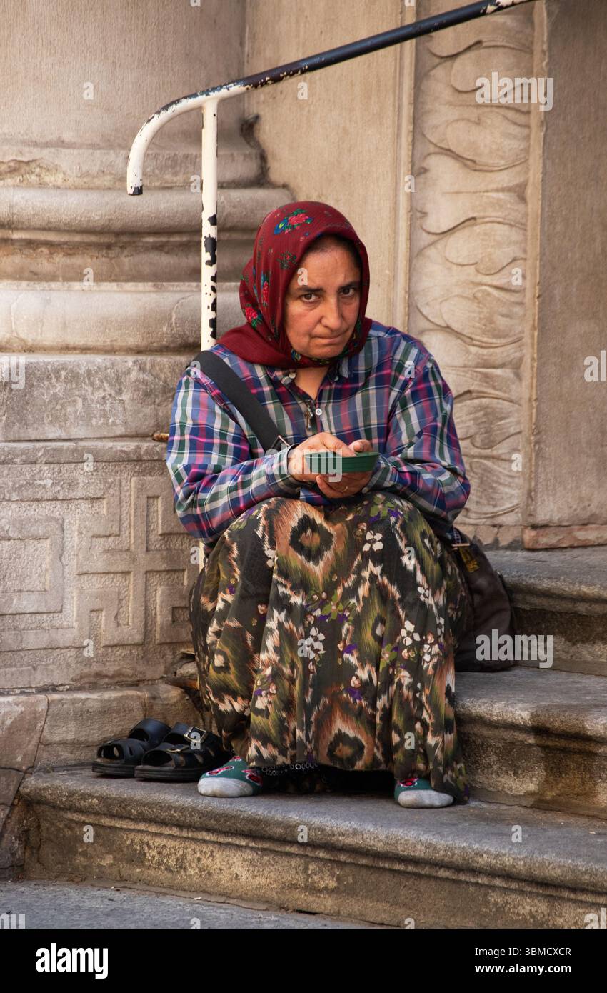 Anziana zingara che implorava da sola in strada seduta tranquillamente in abiti a fantasia concetto di povertà catturato in ambiente urbano emozioni umane grezze Foto Stock
