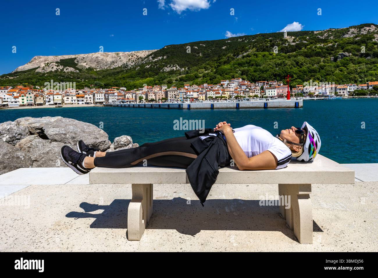 Stanca ciclista si riposa su un prato di cemento nel centro di Baska sull'isola DI KRK, una donna con un casco da bicicletta dorme su una panchina Foto Stock