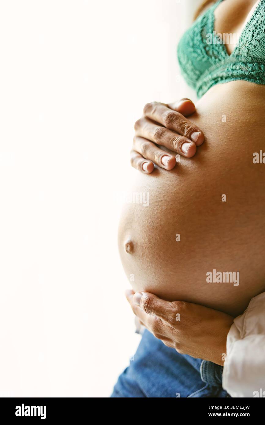 Vista dettagliata della pancia di una mamma incinta cullata da entrambe le mani, con un reggiseno in pizzo verde e jeans, delicatamente illuminata dalla luce della finestra in un tranquillo indoo Foto Stock