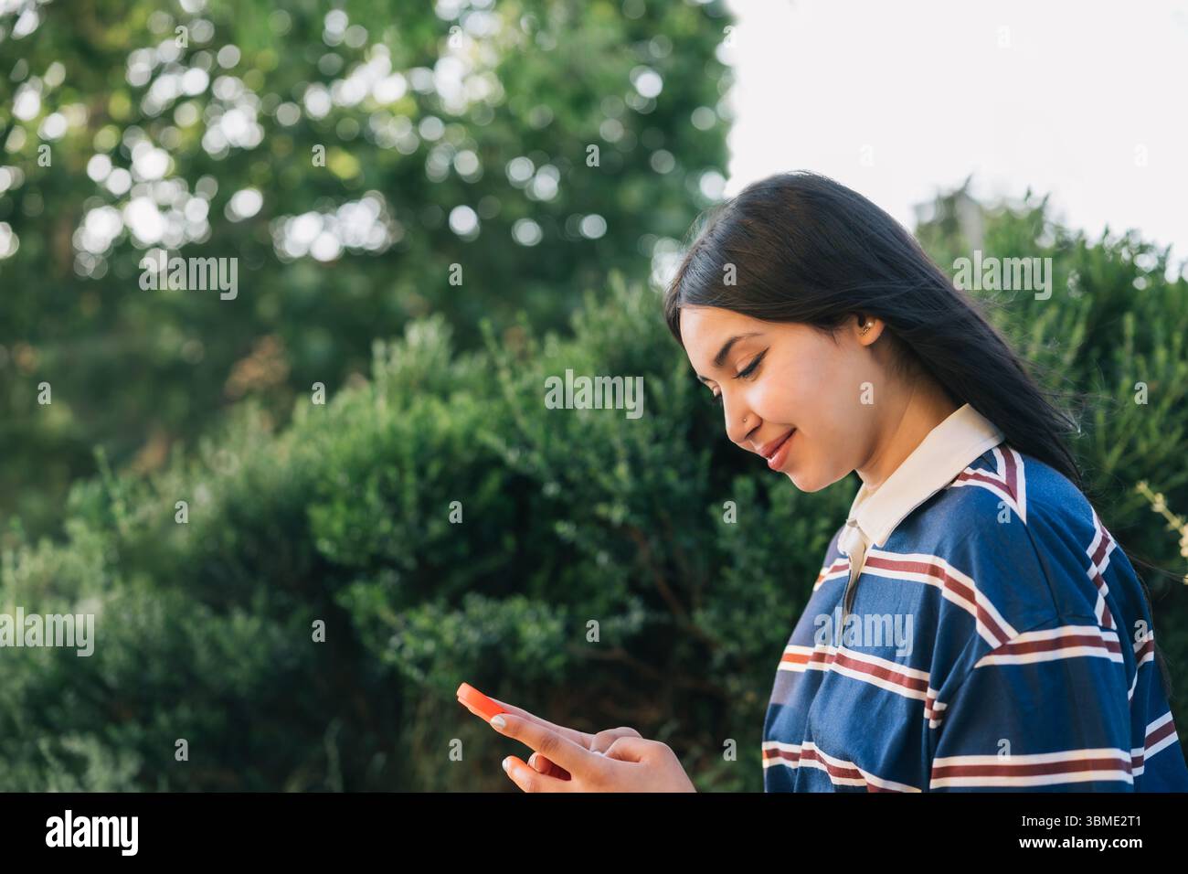 Giovane donna felice con una polo a righe guarda il telefono mentre si gode un momento di tranquillità in un parco verde lussureggiante in una giornata di sole. Stile di vita. Foto Stock