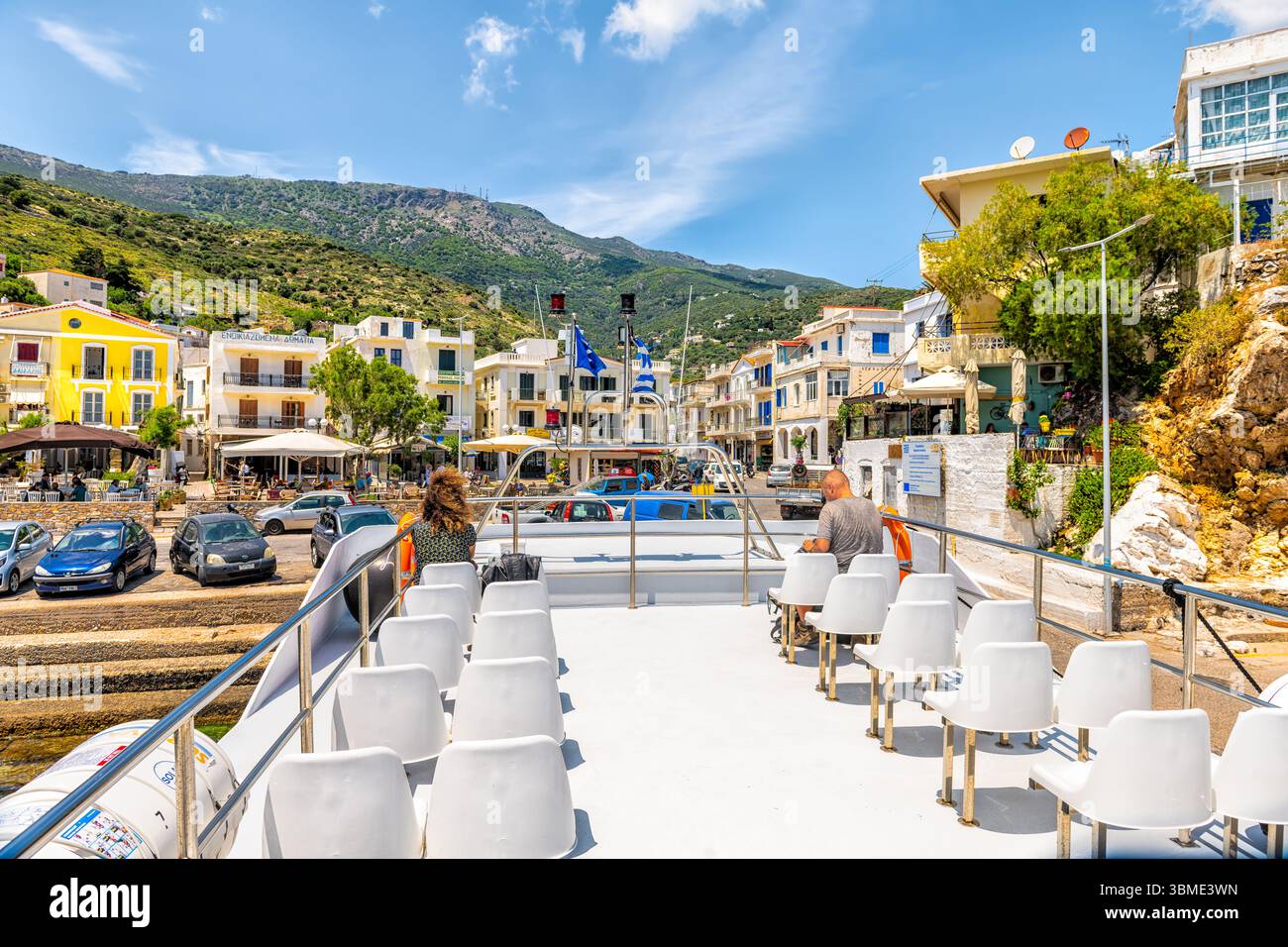 Agios Kirykos, Grecia - 24 maggio 2023: Persone sedute fuori dal ponte dei traghetti con il paesaggio urbano di Agios Kirykos, capitale dell'isola Egea Foto Stock
