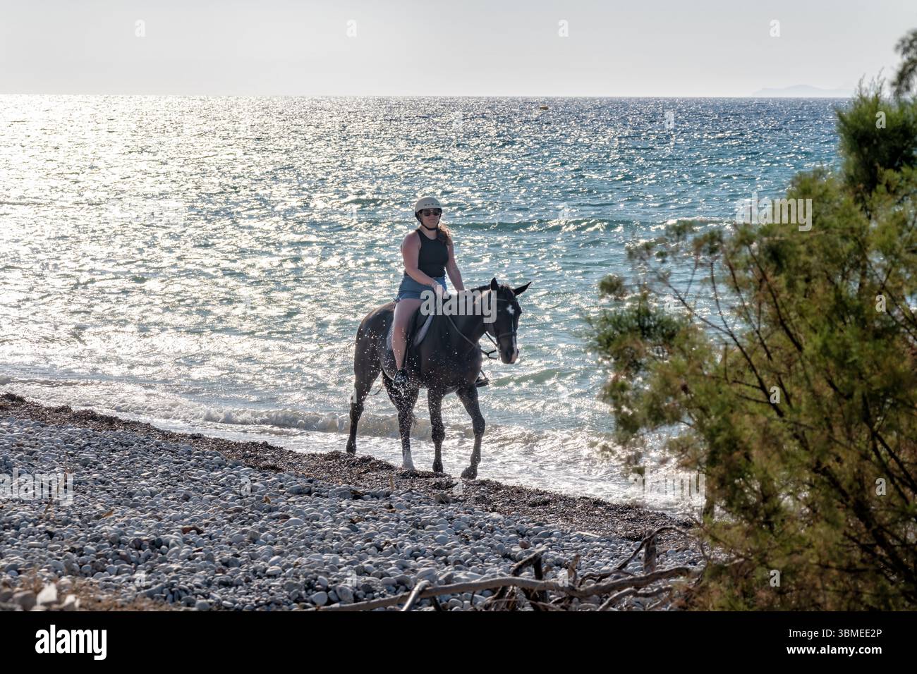 Rodi, Grecia. Una donna turistica cavalca un cavallo lungo una spiaggia. il cavallo cammina vicino al litorale. Un'escursione a cavallo organizzata Foto Stock
