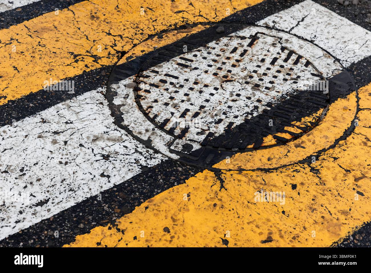 Scena urbana astratta caratterizzata da una strada in asfalto invecchiata con strisce dipinte di giallo e bianco che si intersecano con un tombino di metallo Foto Stock