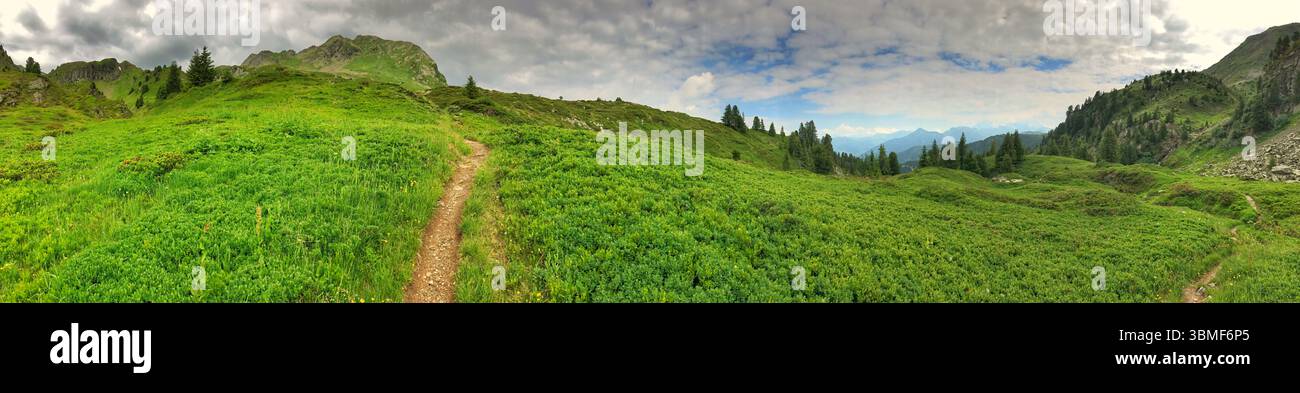 Una fotografia panoramica che mostra un paesaggio verde vivace con colline, sentieri, alberi e montagne lontane, il tutto sotto un cielo blu parzialmente nuvoloso Foto Stock