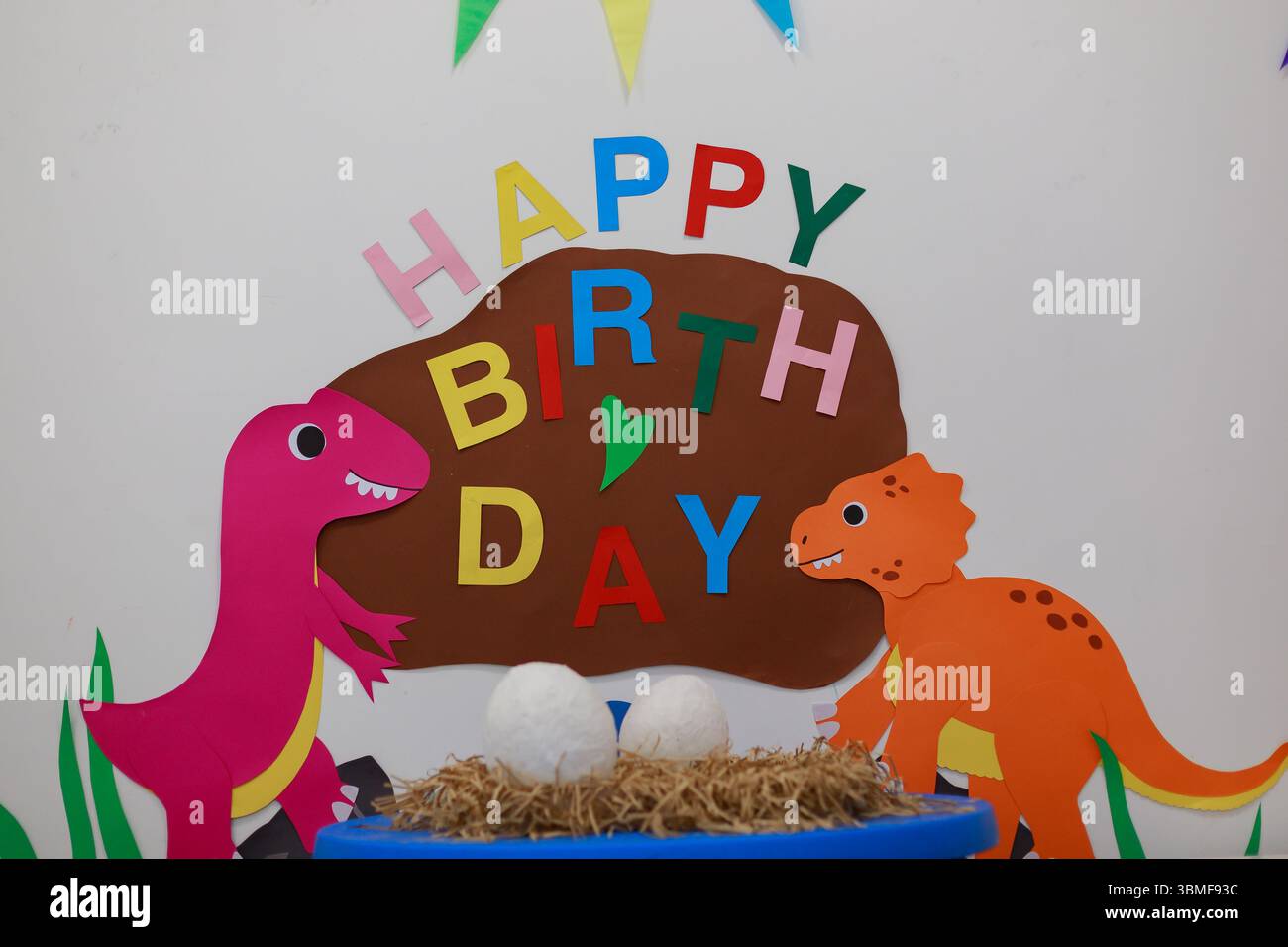Festa di compleanno a tema dinosauro sullo sfondo con lettere DI BUON COMPLEANNO, dinosauri ritagliati in carta, nido di paglia con due uova bianche in primo piano, colorato bu Foto Stock