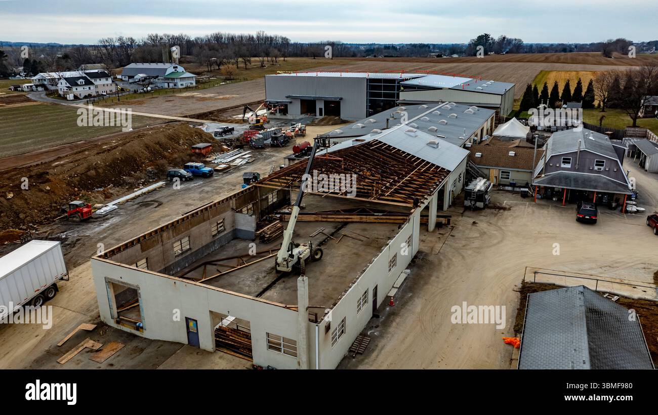 I lavoratori edili gestiscono macchinari in un cantiere in un'area rurale. Le attrezzature pesanti sono visibili mentre metà di una struttura è in fase di ristrutturazione Foto Stock