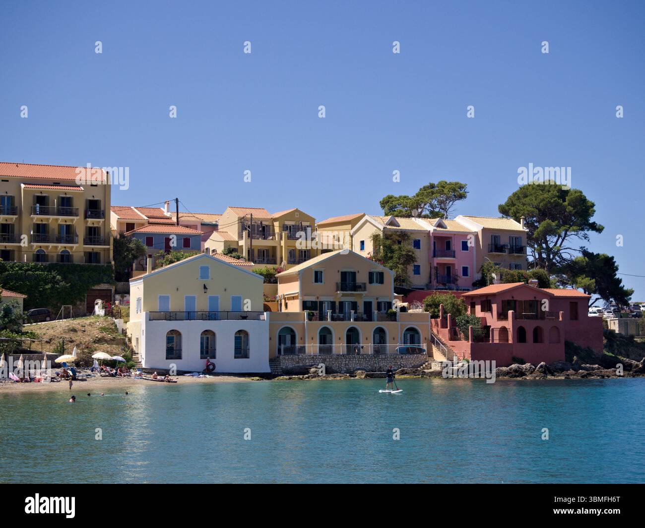 Case colorate del villaggio di Assos, Cefalonia, affacciate sulla baia turchese con alberi di pino e cielo estivo blu. Foto Stock