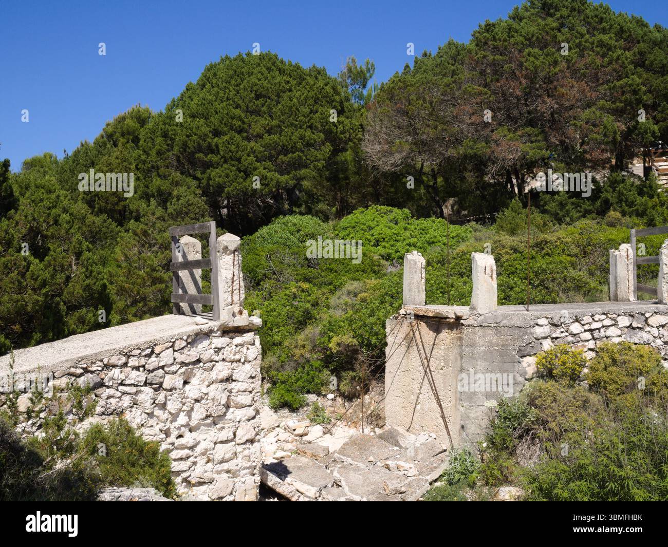 Ponte di pietra bianca rotto che attraversa un letto di fiume secco in un paesaggio mediterraneo con alberi di pino e vegetazione estiva. Foto Stock