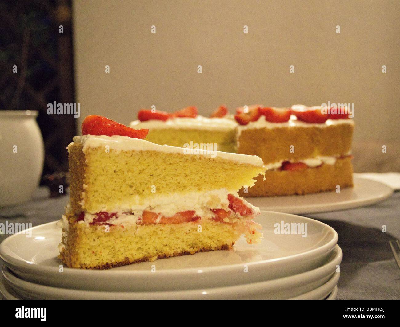 Primo piano della torta di fragole a fette con panna montata in primo piano e la torta completa visibile sullo sfondo. Foto Stock