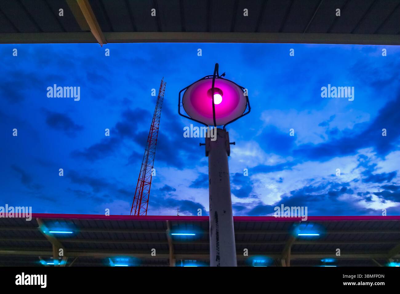 Scatto ad angolo basso della lampada rosa neon e della torre delle telecomunicazioni al crepuscolo. Il cielo drammatico aggiunge un moderno umore urbano cyberpunk e la scena dei trasporti notturni. Foto Stock