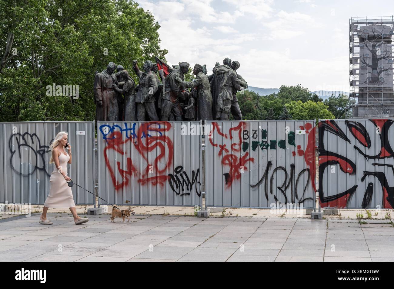Sofia, Bulgaria. 21 giugno 2025. Una donna cammina il suo cane davanti al monumento dell'esercito sovietico, parzialmente smantellato e imbarcato, costruito nel centro di Sofia Foto Stock