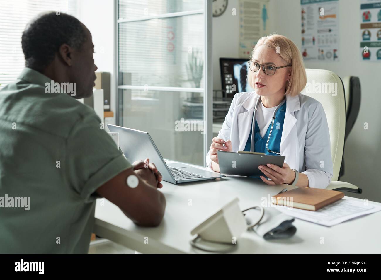 Donna caucasica di mezza età medico consulenza uomo nero di mezza età con diabete in studio medico, medico che tiene appunti e penna, paziente wearin Foto Stock