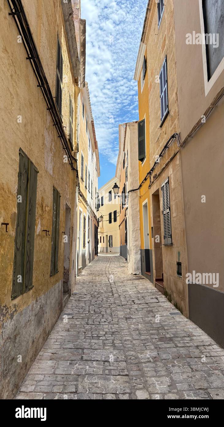 Stretta strada di ciottoli dall'estetica con edifici gialli e beige a Minorca, Spagna durante l'estate. - Immagine stock catturata con smartphone