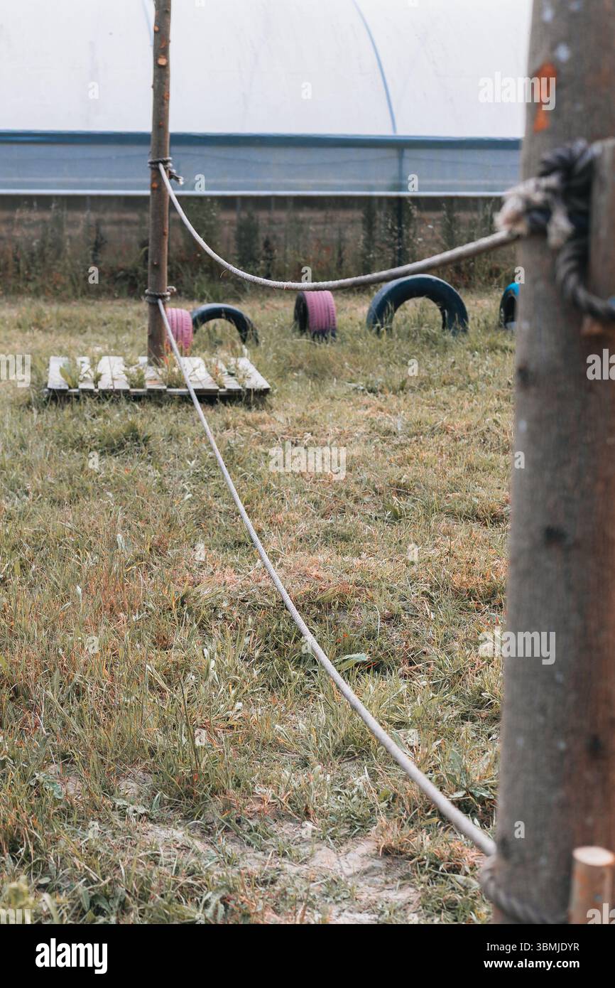 Primo piano di un ostacolo di corda in un'area giochi all'aperto per bambini con erba, pneumatici ed elementi di legno, davanti a una struttura a serra Foto Stock