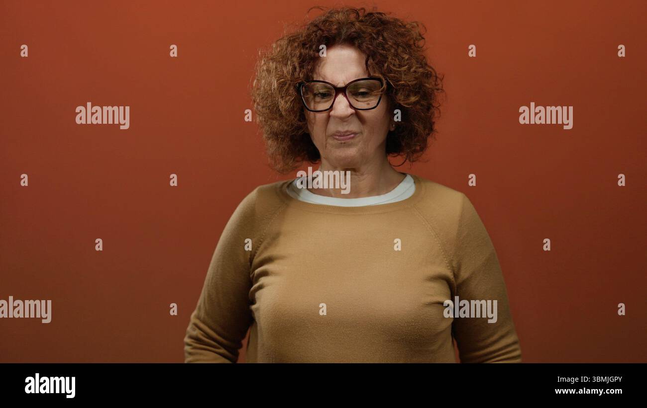 Una donna ispanica di mezza età con capelli ricci fa uno strano volto reagendo a un cattivo odore su uno sfondo marrone. Foto Stock