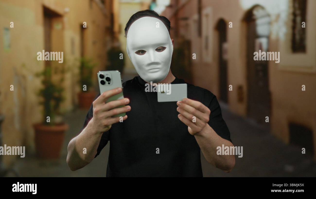 Un giovane in maschera tiene il telefono e la carta sulla strada della città, simboleggiando la sicurezza digitale e il furto di identità. Foto Stock